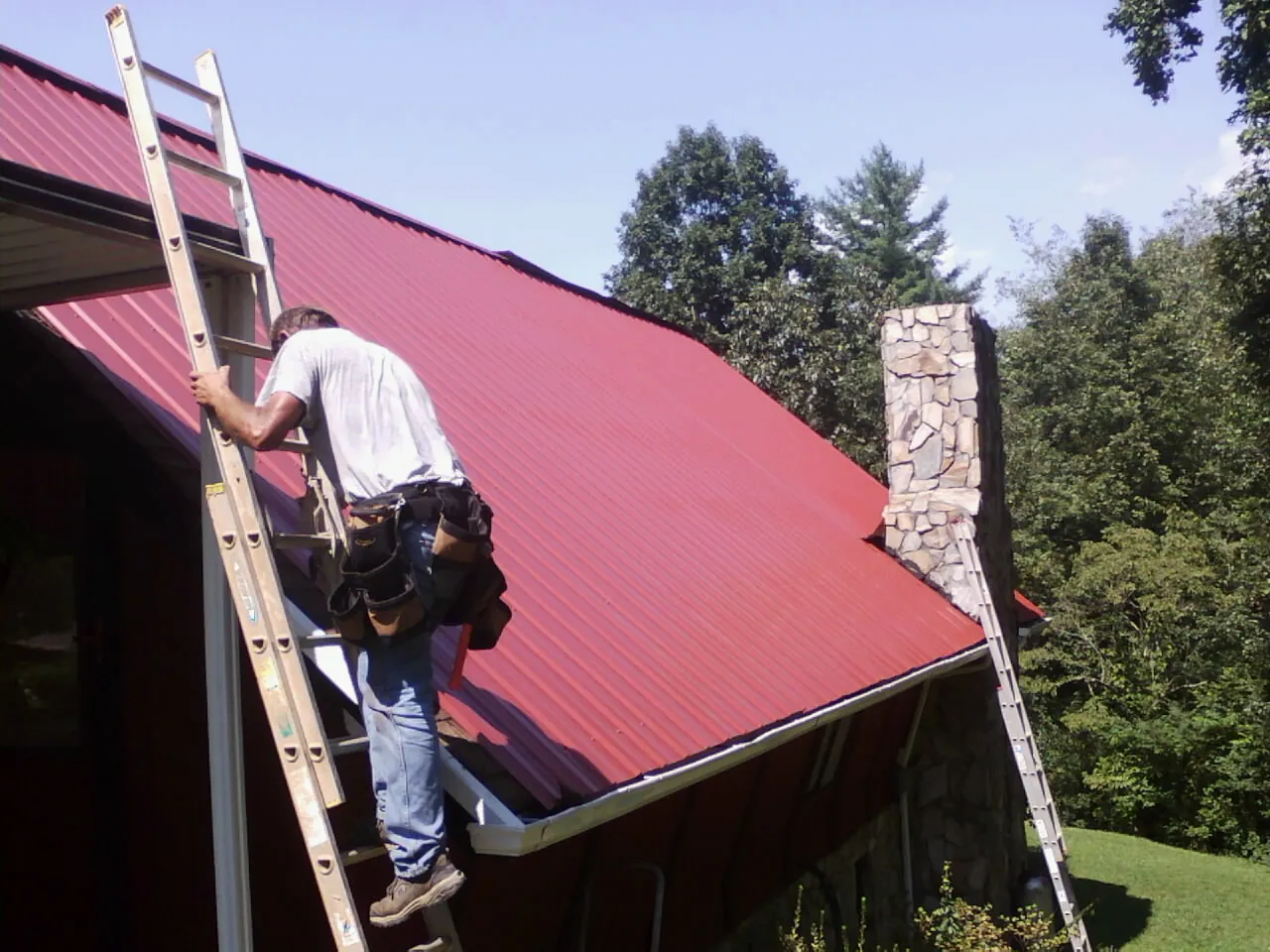 Roofer on a ladder beside a red metal roof and stone chimney — Soffit & Fascia Repair by Roofing Brothers in Chalfont